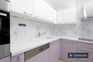 Svetla bela kuhinja z pastelno vijoličastimi omaricami in belim kuhinjskim steklom Luminum na katerem so regratove lučke v nežni vijola barvi, enaki barvam kuhinjkih frontov